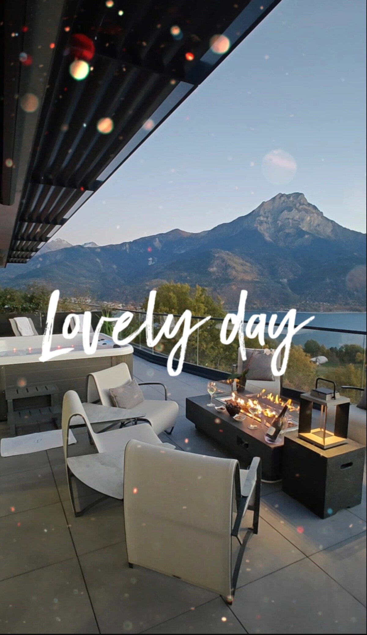 An outdoor terrace is displayed, featuring modern white seating and a fire pit. Scenic mountain views are visible in the background, with a lake reflecting the surrounding landscape. Soft lighting creates an inviting atmosphere while the natural surroundings enhance the overall environment.