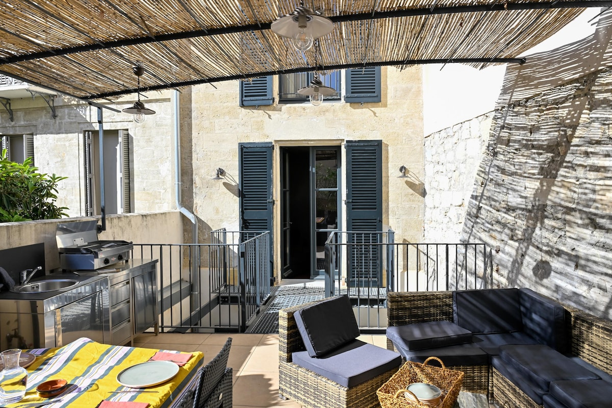 A private terrace is featured with a woven shade structure providing soft light. Comfortable seating is arranged with dark cushions, and a dining area is set with a colorful tablecloth. Double doors lead to the interior space, enhancing the inviting outdoor atmosphere.