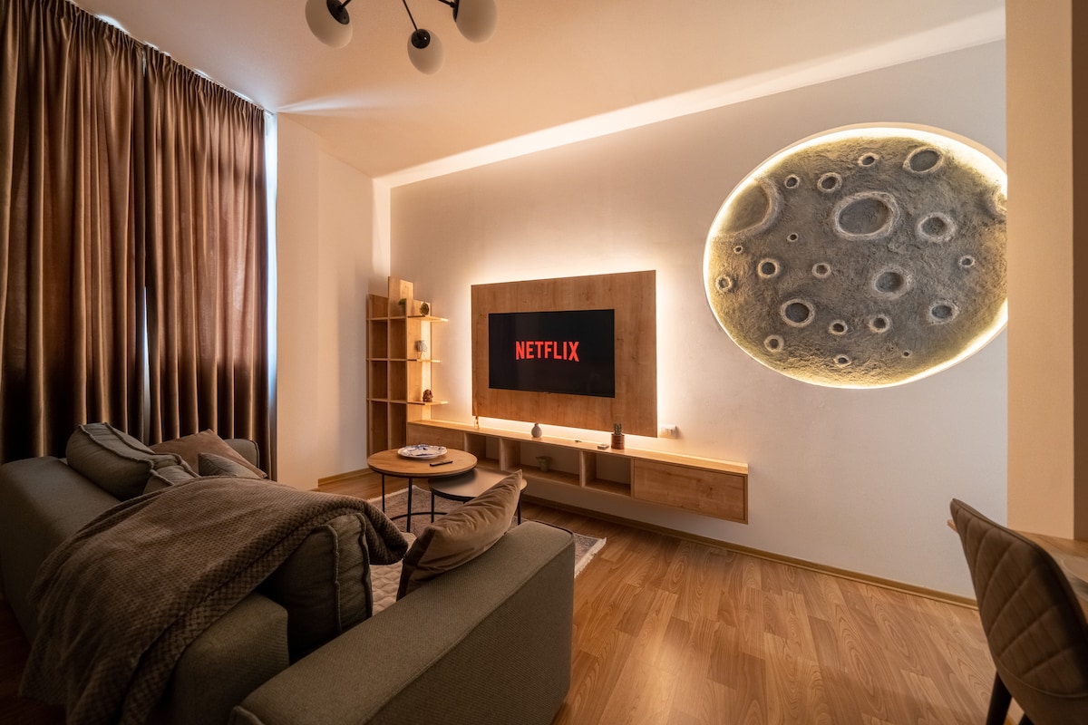 A modern living area features a soft gray sofa with an inviting throw blanket. A wall-mounted television displays a streaming interface. Warm lighting accents a large, decorative wall feature resembling the moon, while light drapes frame a large window, enhancing the airy feel of the space.