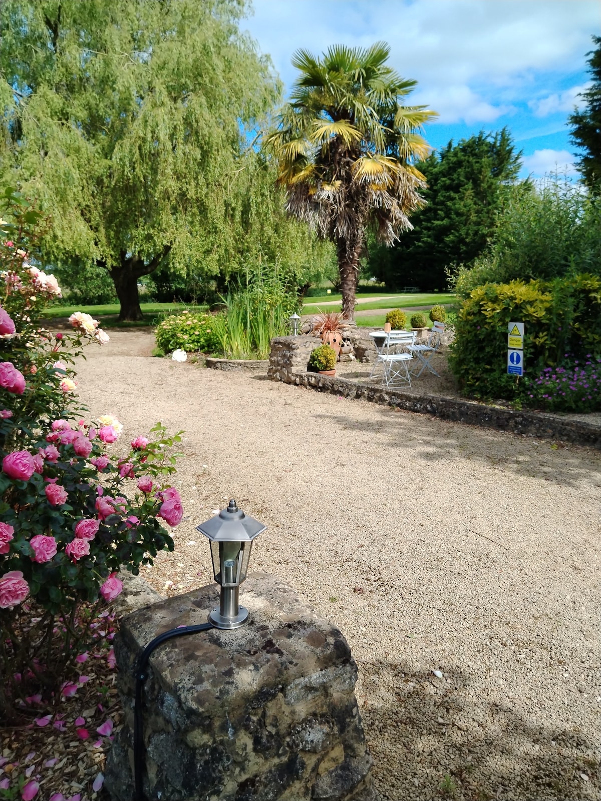 A serene garden path is framed by blooming rose bushes and lush greenery, with a decorative lantern placed on a stone pillar. In the background, a palm tree and various seating areas are visible amidst the well-maintained landscaping.