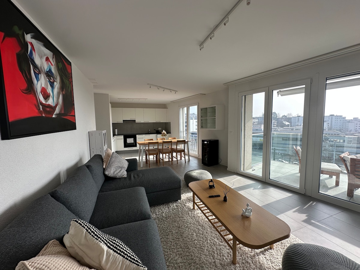 A spacious living room features a large grey sectional sofa accompanied by a wooden coffee table. Light streams in through floor-to-ceiling windows that lead to a balcony, showcasing city views. A dining table with several chairs and a vibrant piece of wall art adds character to the space.