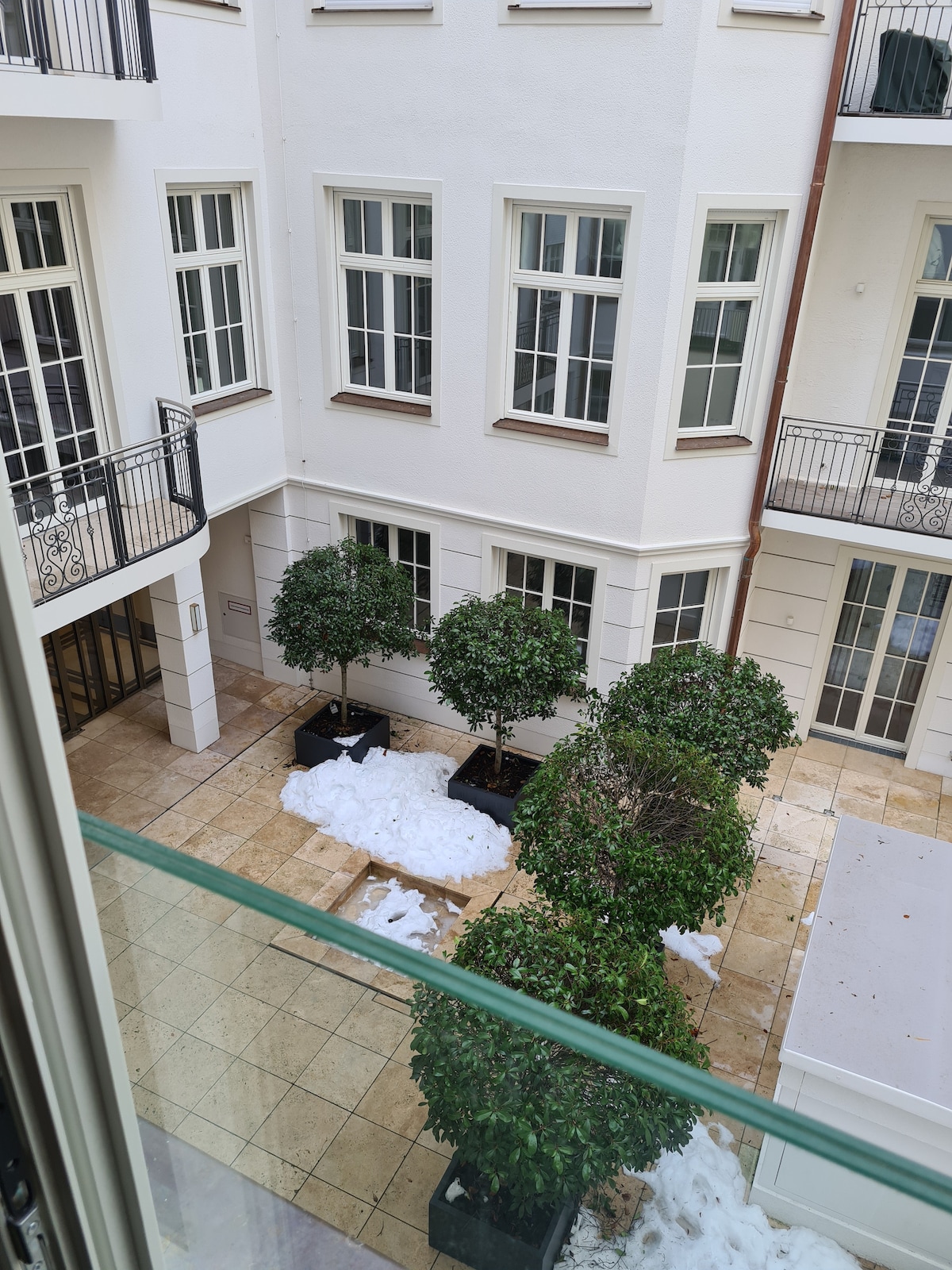 A view into a serene courtyard features neatly trimmed bushes housed in black planters. Snow remnants rest on the tiled ground, surrounded by white walls and large windows of the building. Balconies with decorative railings provide an elegant architectural detail.