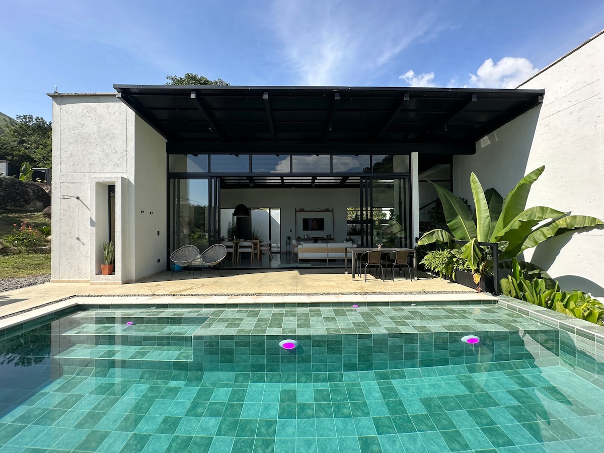 An inviting outdoor space features a sparkling pool bordered by turquoise tiles. A shaded patio provides seating areas, complemented by tropical plants. Large glass doors open up to the interior, creating a seamless connection between indoor and outdoor spaces.