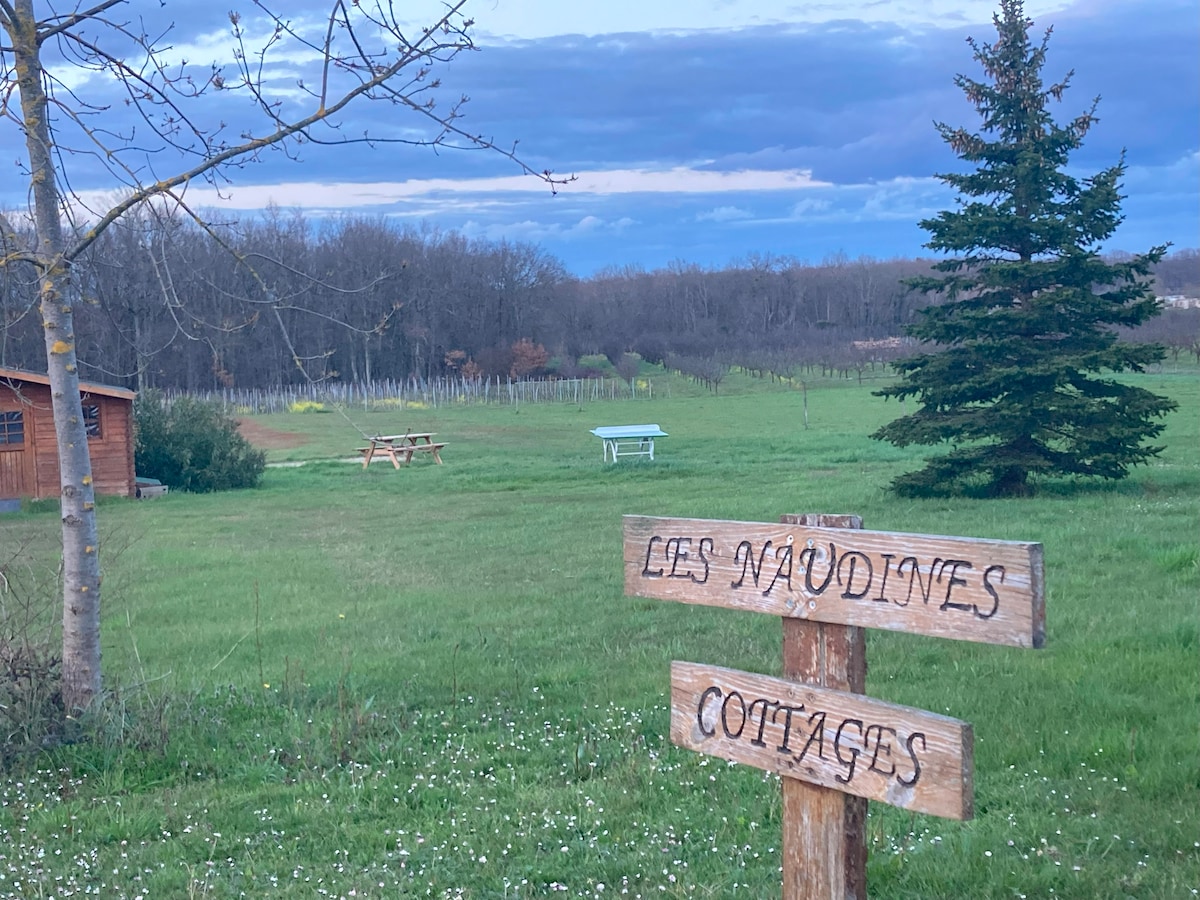 A wooden sign marks the entrance to Les Naudines Cottages, surrounded by lush green grass and small wildflowers. In the background, a mix of trees and a tranquil landscape can be seen under a softly clouded sky, enhancing the natural setting.