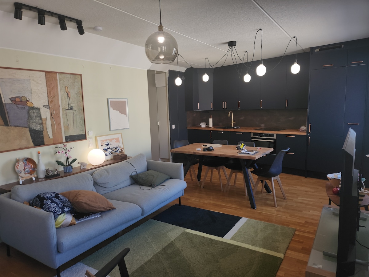 A spacious living room features a light grey sofa arranged alongside a green and blue area rug. A wooden dining table is accompanied by four chairs and is positioned near a modern kitchen with dark cabinetry. Decorative art pieces adorn the walls, and pendant lights hang above.