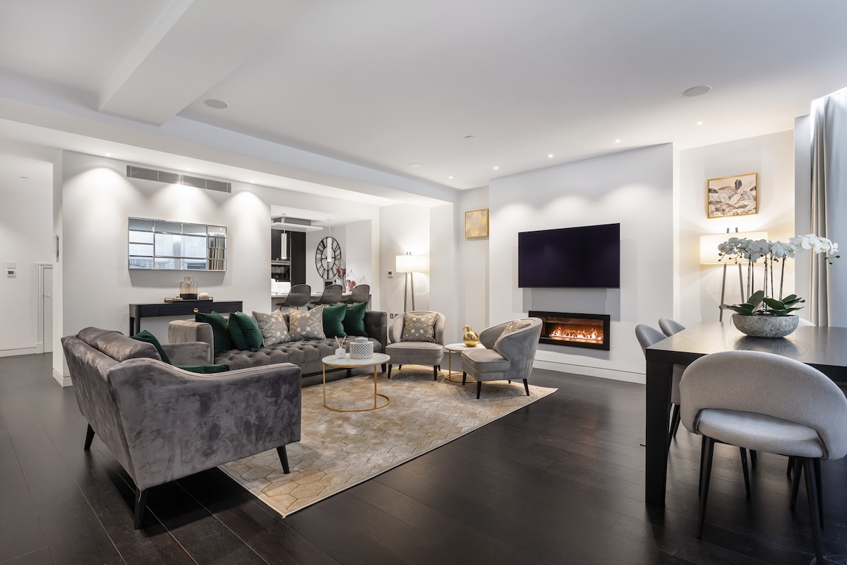 A modern living area features a spacious arrangement of plush seating in muted tones, complemented by a stylish coffee table. A wall-mounted flat-screen TV is positioned above an elegant electric fireplace, while a dining table is set for six, illuminated by soft lighting.