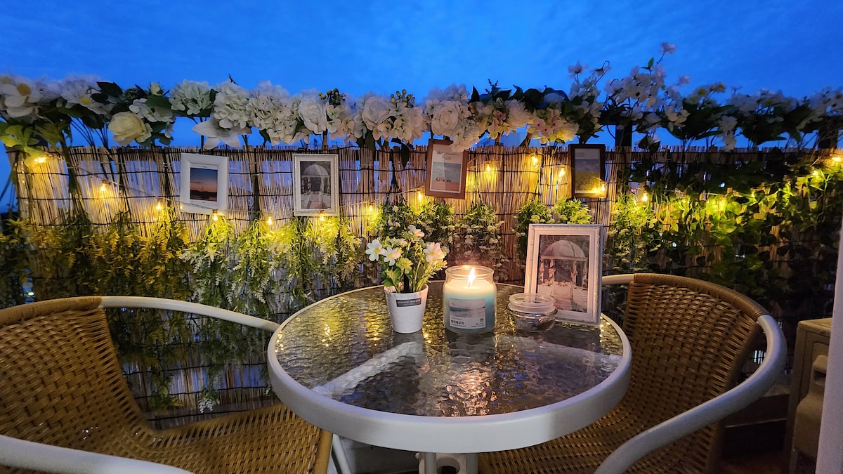 An outdoor terrace is adorned with decorative flowers and soft lighting. A circular glass table is surrounded by two wicker chairs. A lit candle sits atop the table next to a small potted plant and framed photographs, creating a serene and inviting atmosphere.