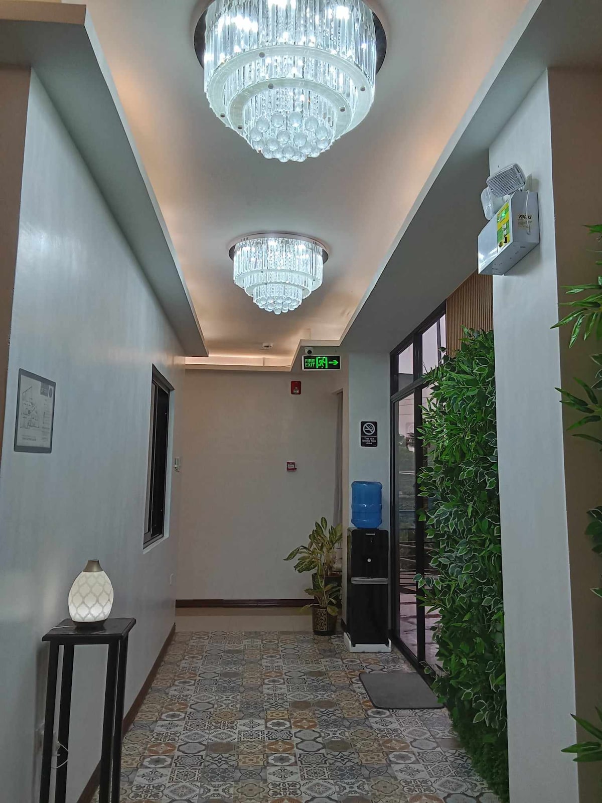 A well-lit hallway features three modern chandeliers hanging from the ceiling. Decorative tiles line the floor, and a small table with a lamp stands beside a lush green plant. Natural light filters through a large window at the end of the corridor.
