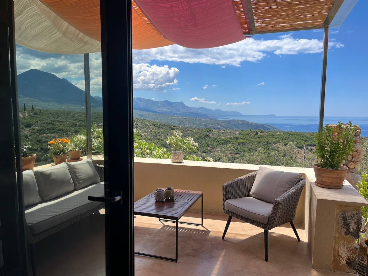 A shaded outdoor terrace offers a comfortable seating area with a sofa and chairs, enhanced by potted plants. Scenic views of rolling hills and the sea can be seen in the background, under a blue sky filled with clouds.