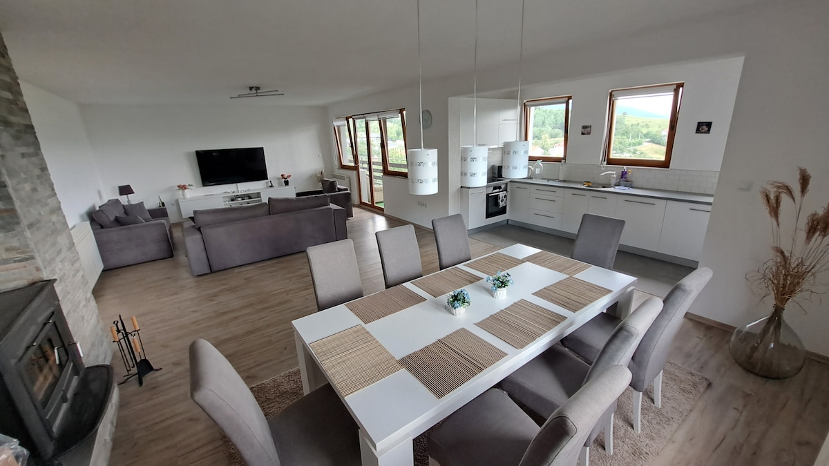 A spacious living area is showcased, featuring a modern kitchen and dining area with a large table surrounded by grey upholstered chairs. The room is filled with natural light from multiple large windows, offering views of the surrounding landscape. A comfortable sitting area is positioned behind.