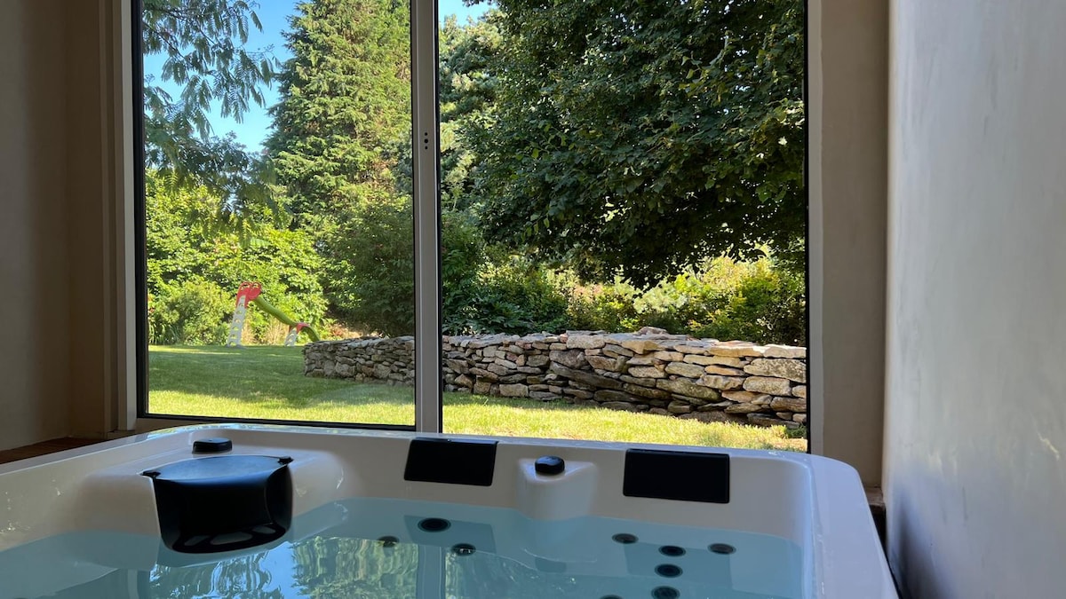 A hot tub is positioned beside a large window, providing a view of a lush garden with greenery and a stone wall. Natural light fills the space, enhancing the calming atmosphere.