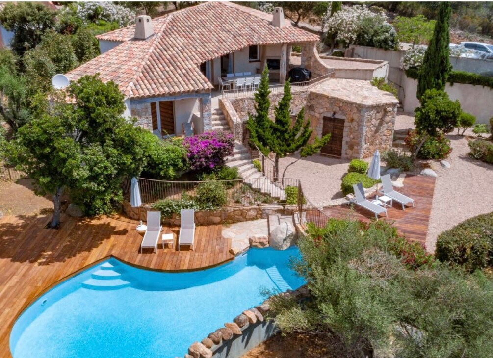 A charming stone villa is surrounded by lush greenery and colorful flowers. A clear pool is set within a wooden deck area, complemented by several sun loungers. Steps lead to a cozy outdoor dining space, enhancing the inviting atmosphere of the property.