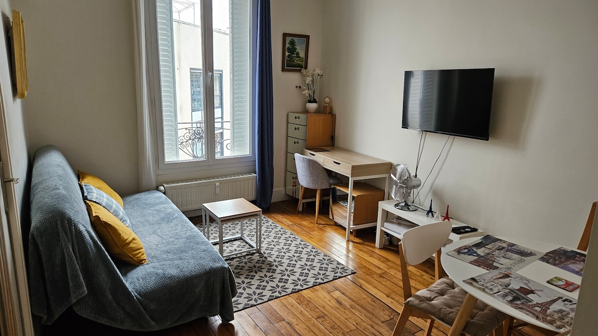 The living area features a comfortable sofa bed dressed in soft gray fabric, accompanied by decorative yellow cushions. A light wooden table and chairs for dining are positioned near a window, with a mounted TV and a modern desk against the wall. A patterned rug enhances the warm wooden flooring.