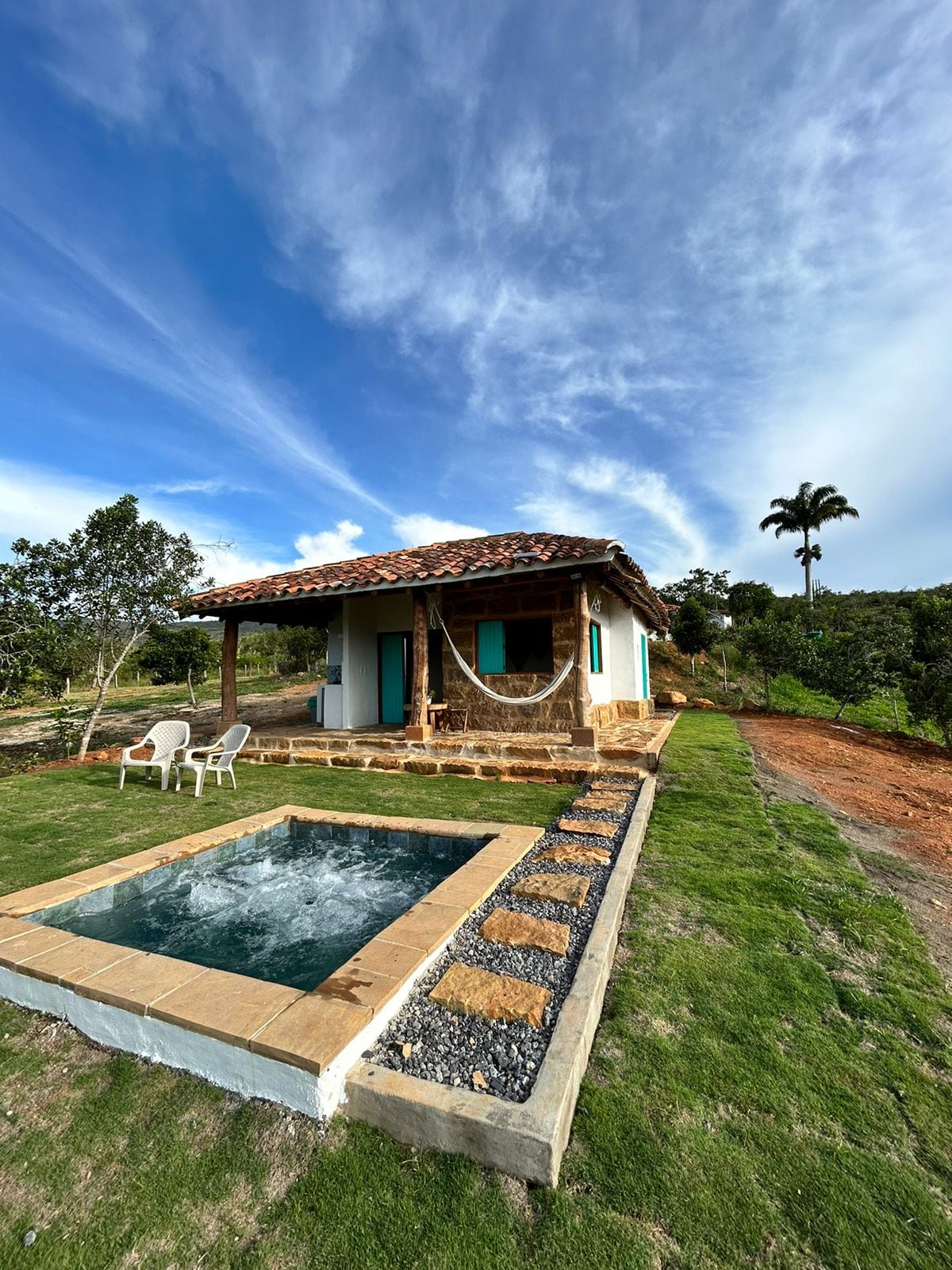 A charming colonial-style cabin sits amidst natural greenery, featuring a private jacuzzi beside it. The structure features large windows with blue shutters and is topped with a traditional tiled roof. Comfortable seating is placed beside the jacuzzi, inviting relaxation under a clear sky.