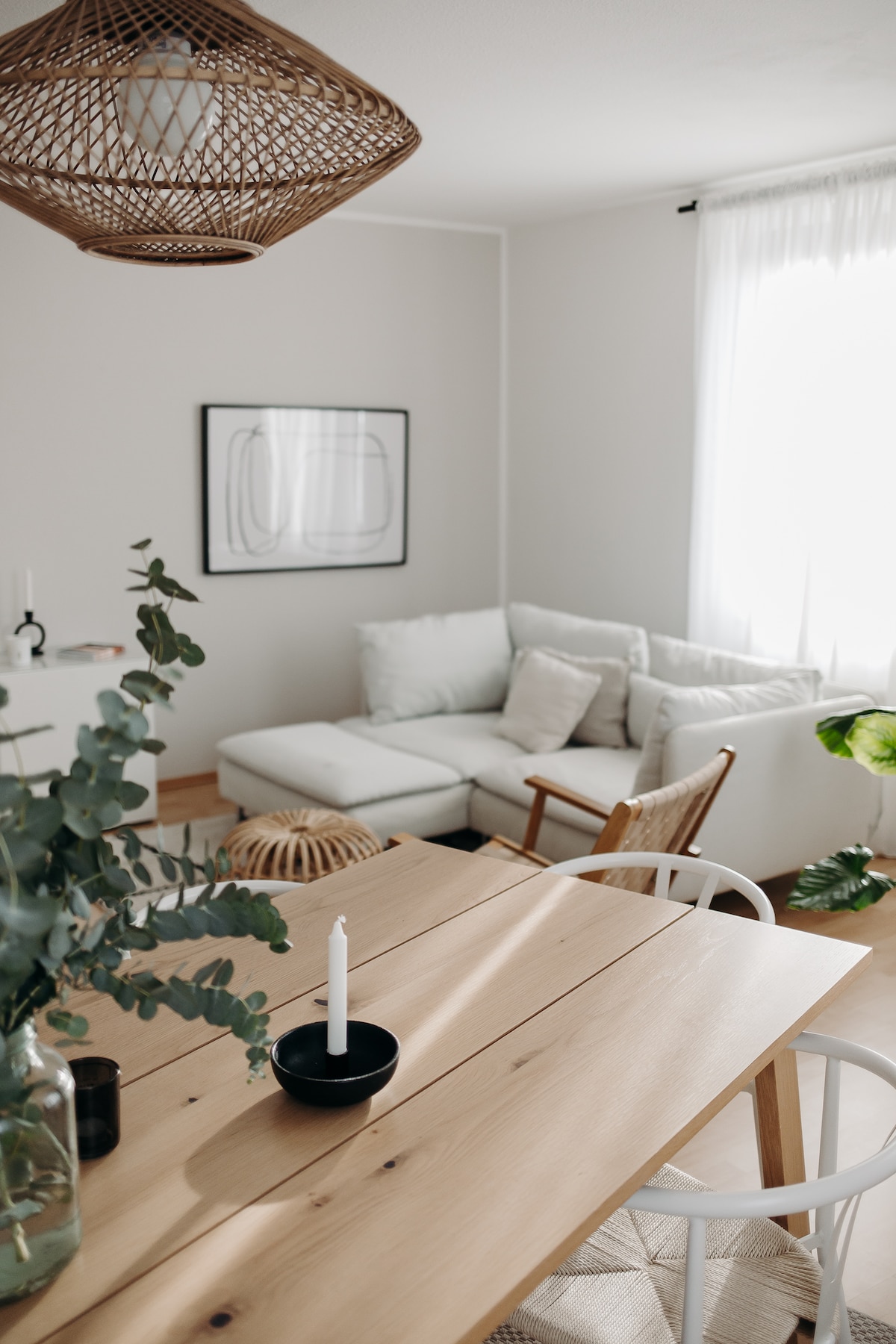 A cozy living area features a light-colored sectional sofa, enhanced by decorative pillows. A wooden dining table is set with a candle and a small vase, while greenery adds a natural touch. Soft natural light filters through a sheer curtain, highlighting the room's warm atmosphere.