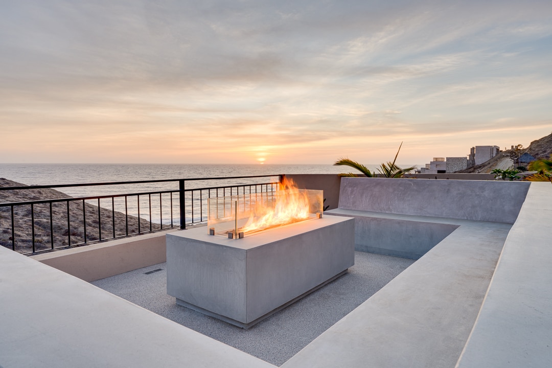 A modern outdoor space features a sleek fire pit surrounded by seating, set against a backdrop of the ocean and a vibrant sunset. Soft colors in the sky enhance the tranquil atmosphere, providing an inviting setting for relaxation.