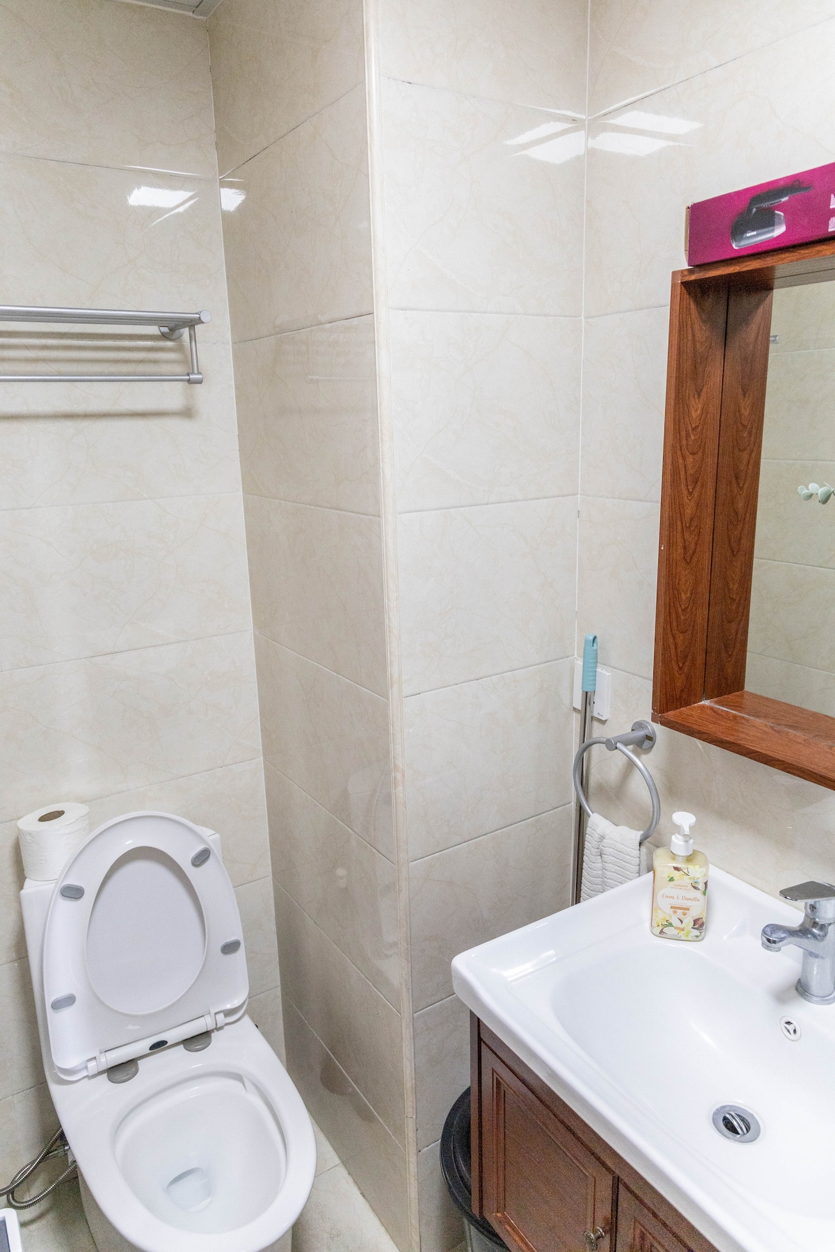 A clean bathroom space is featured with tiled walls and a large mirror above the sink. A wooden cabinet provides storage beneath the sink. Essentials like soap are present, and a towel bar is mounted on the wall. A toilet is positioned for accessibility.