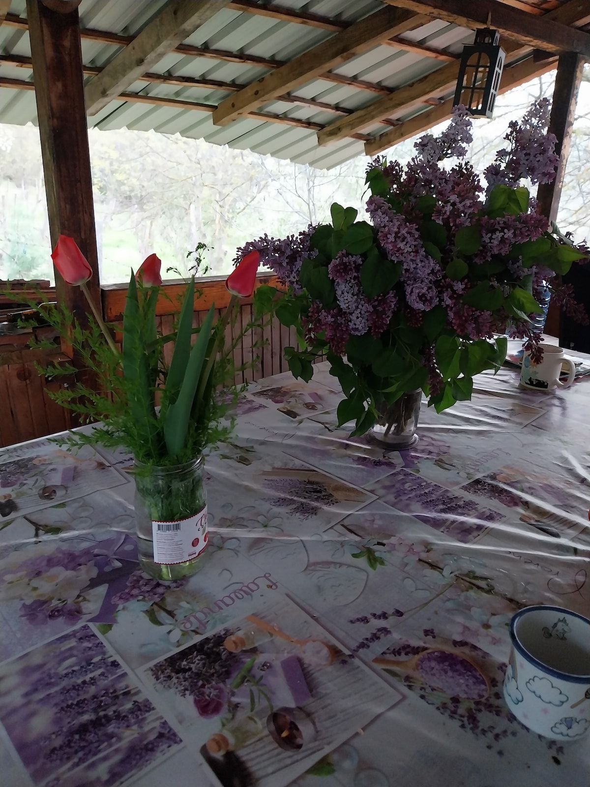 A rustic wooden table is adorned with a floral tablecloth and features two arrangements. One includes tulips and green foliage in a glass jar, while the other showcases a large bouquet of lilacs in a vase. Natural light filters in from the surrounding windows.