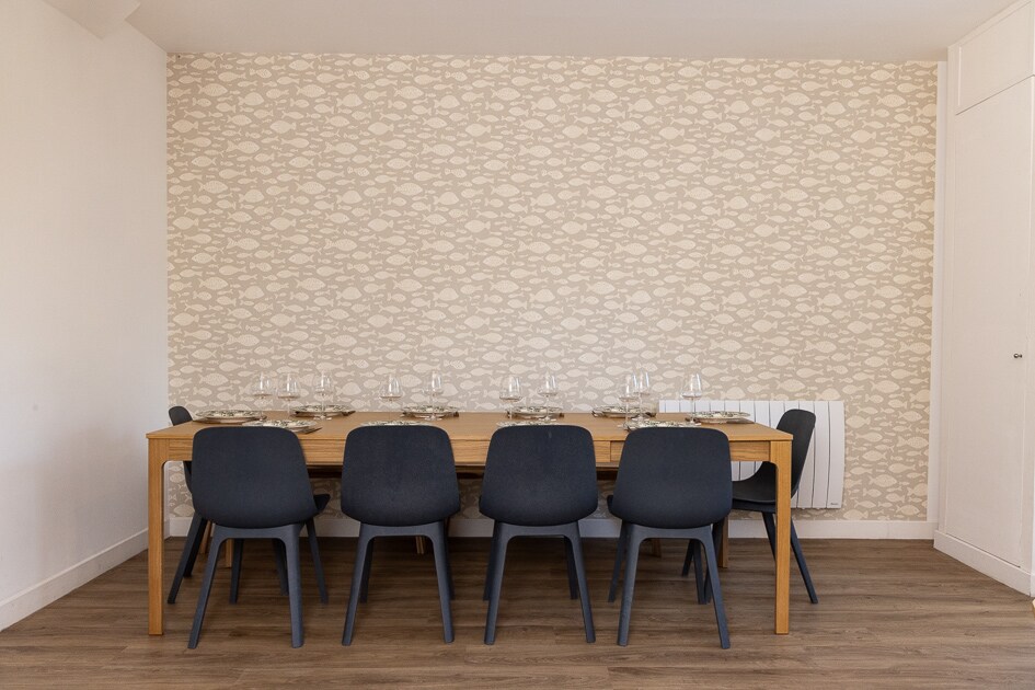 A dining area is presented with a large wooden table, designed to accommodate multiple guests. Dark chairs surround the table, which is set with clear glassware. The backdrop features a softly patterned wall, complementing the natural tones of the floor.