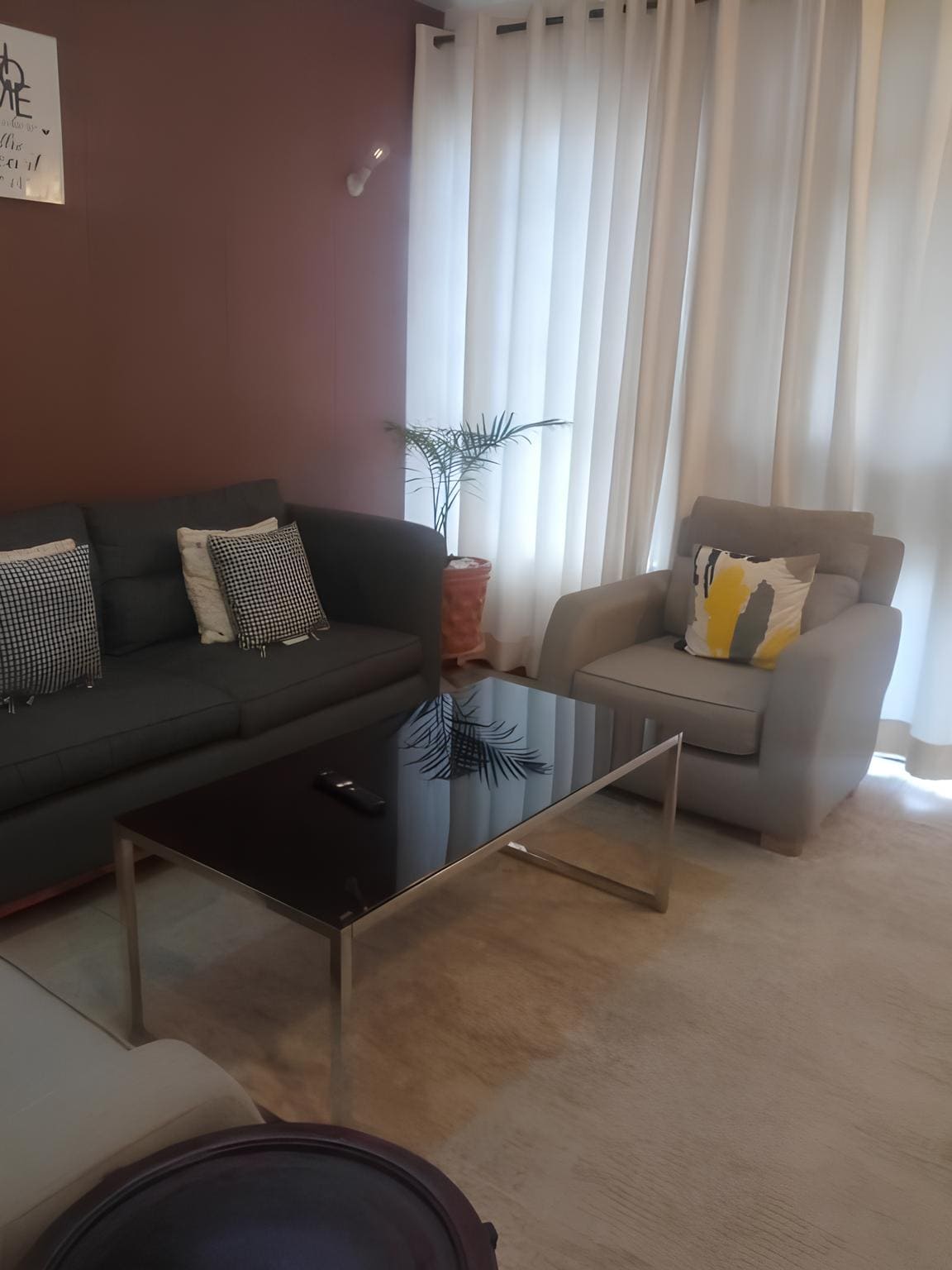 A cozy living area features a comfortable gray sofa paired with a light gray armchair. A glass coffee table sits in the center, reflecting natural light that filters through the sheer curtains. Decorative pillows add a subtle touch of color, enhancing the space's relaxing atmosphere.