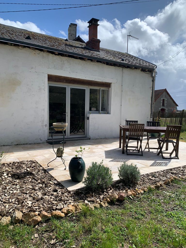 An outdoor patio features a wooden dining set with four chairs and a small table, complemented by decorative planters. A barbecue grill is positioned nearby. The area is surrounded by landscaped gravel and green grass, with a backdrop of the gite's exterior and a partly cloudy sky.