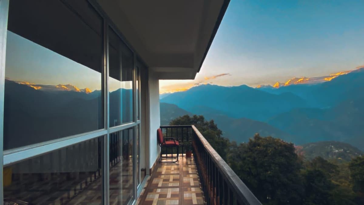 A balcony offers a stunning view of surrounding mountains, with a single chair positioned to take in the scenery. Large glass doors reflect the landscape, creating a sense of openness as evening light bathes the area.