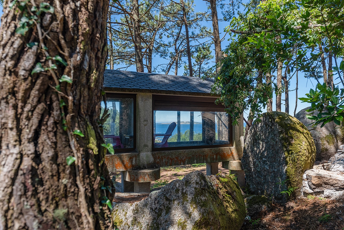 A modern structure is nestled among trees and rocks, featuring large windows that provide a view of the ocean. Sunlight filters through the foliage, creating a serene outdoor setting that emphasizes the natural surroundings.