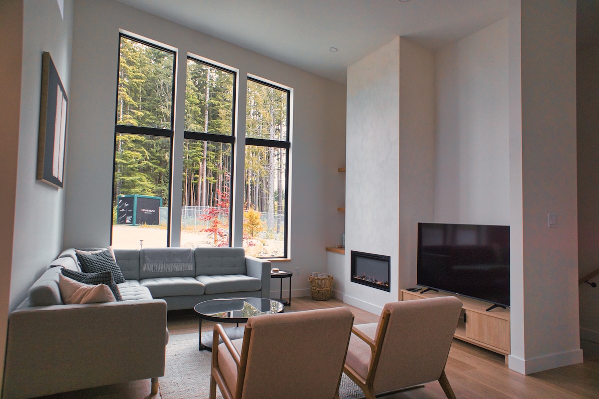 A spacious living area is highlighted by tall windows, providing views of the forest. A plush sectional couch faces a modern television, while two armchairs are positioned nearby. A circular coffee table sits at the center, enhancing the inviting ambiance of the room.