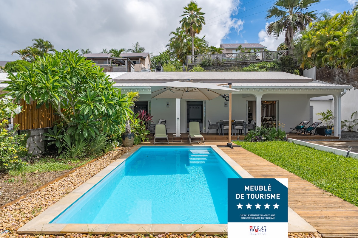 The image displays a modern villa with a clear blue pool surrounded by a wooden deck. Lush tropical plants frame the scene, while a shaded seating area under an umbrella invites relaxation. The structure is characteristic of tropical architecture, offering comfort and style.