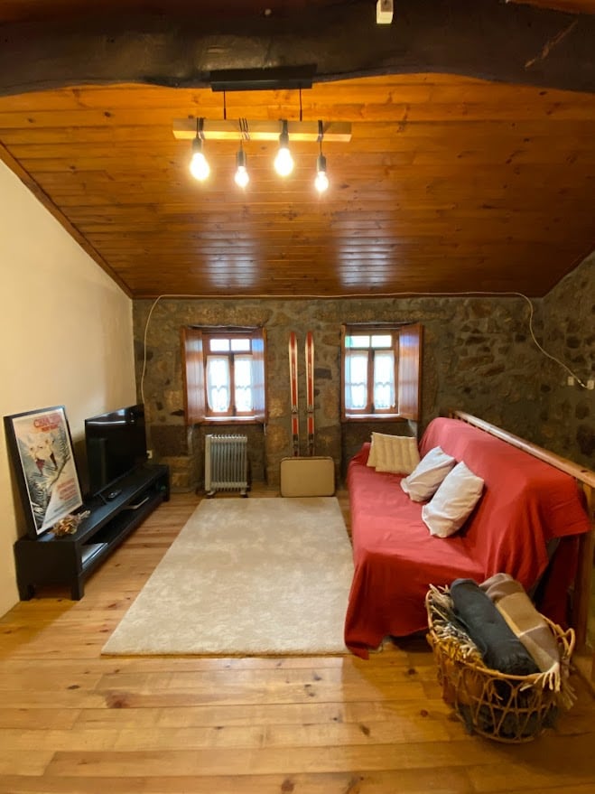 A bright living area features a red sofa adorned with cushions, positioned next to a small basket. A TV unit holds a television, while two windows allow natural light to fill the space. Light fixtures hang from the wooden ceiling above.