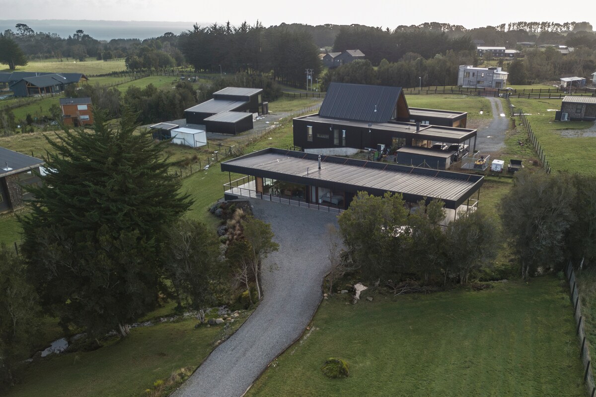 An aerial view captures the spacious outdoor area surrounding a modern black house. A gravel pathway leads to the entrance, bordered by greenery. The property features a mix of buildings, sprawling lawns, and trees under a soft sky, highlighting the serene and expansive landscape.