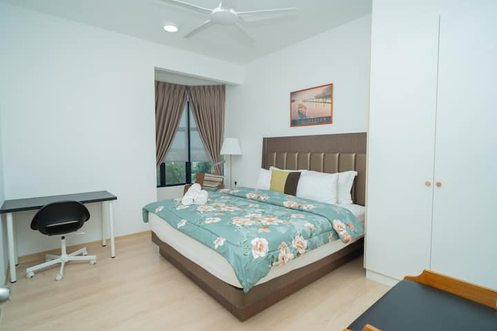 Cozy Bedroom 2 with a queen-sized bed, floral bedding, and a large padded headboard. Features include a study desk, full-height wardrobe, and window with blackout curtains—ideal for work and rest.