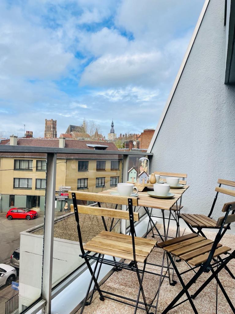 A balcony is depicted with two wooden folding tables and four matching chairs, offering a space for outdoor seating. A city skyline, complete with historic architecture and a steeple, is visible in the background under a partly cloudy sky.
