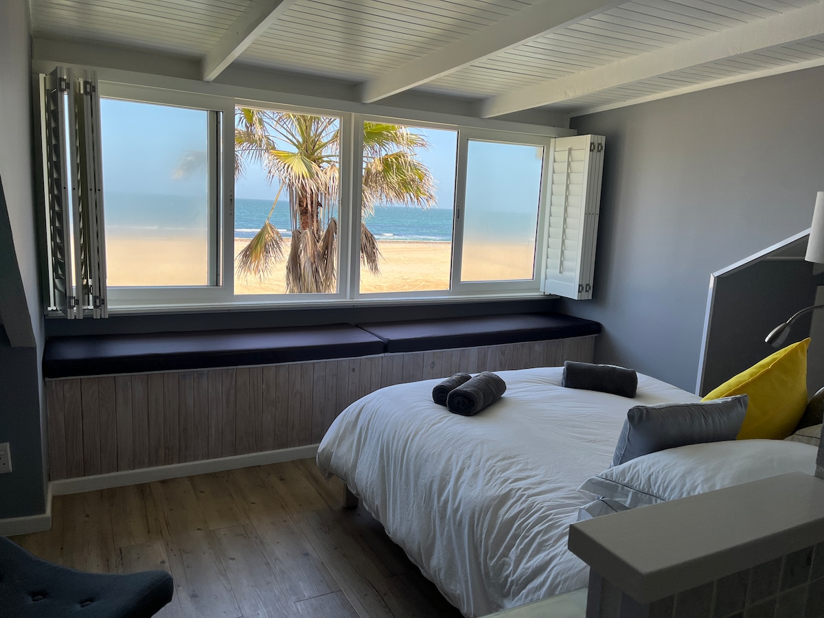 A serene bedroom features a spacious bed dressed in crisp white linens. Large windows showcase a stunning sea view, while a palm tree can be seen on the beach outside. A comfortable bench is positioned beneath the windows, providing additional seating.