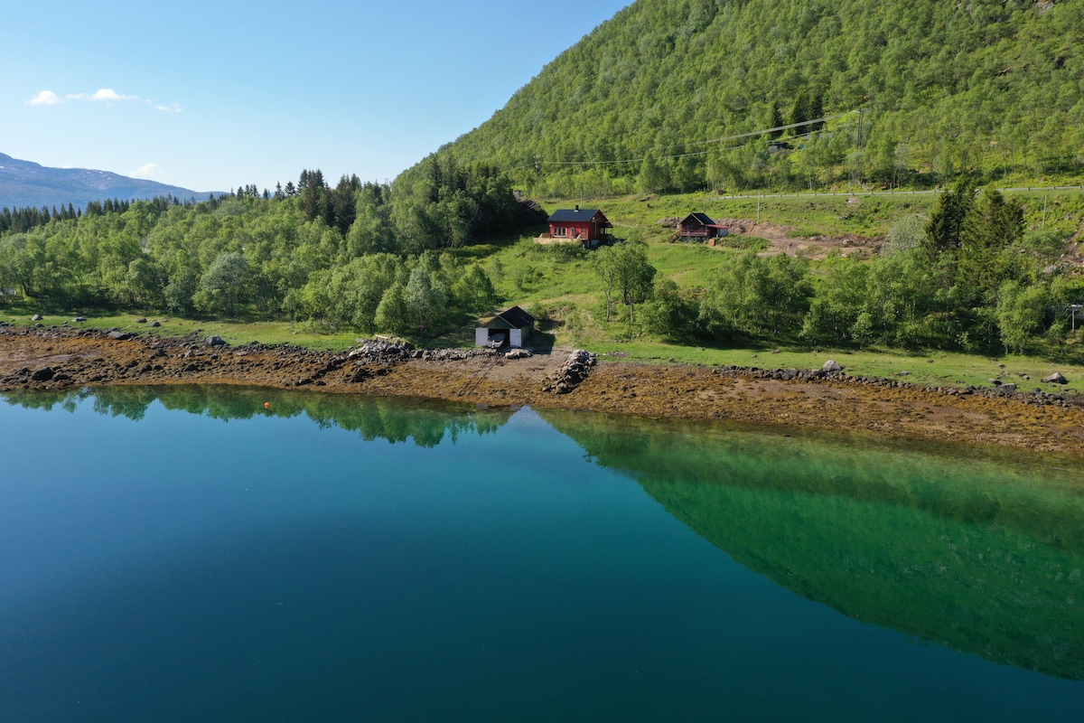 A scenic view of a calm water body reflecting the surrounding greenery and mountains. The shoreline is lined with smooth stones, and a few rustic buildings are visible on the hill, surrounded by lush trees under a clear sky.