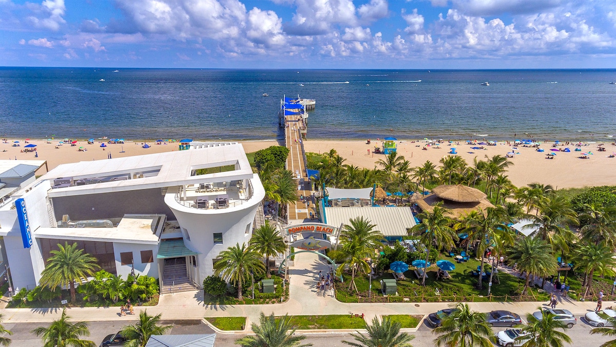 An aerial view captures a beach scene with soft golden sands and gentle waves. The blue water meets the horizon under a partly cloudy sky. A wooden pier extends from the shore, flanked by palm trees and beach umbrellas, creating a relaxed coastal atmosphere.