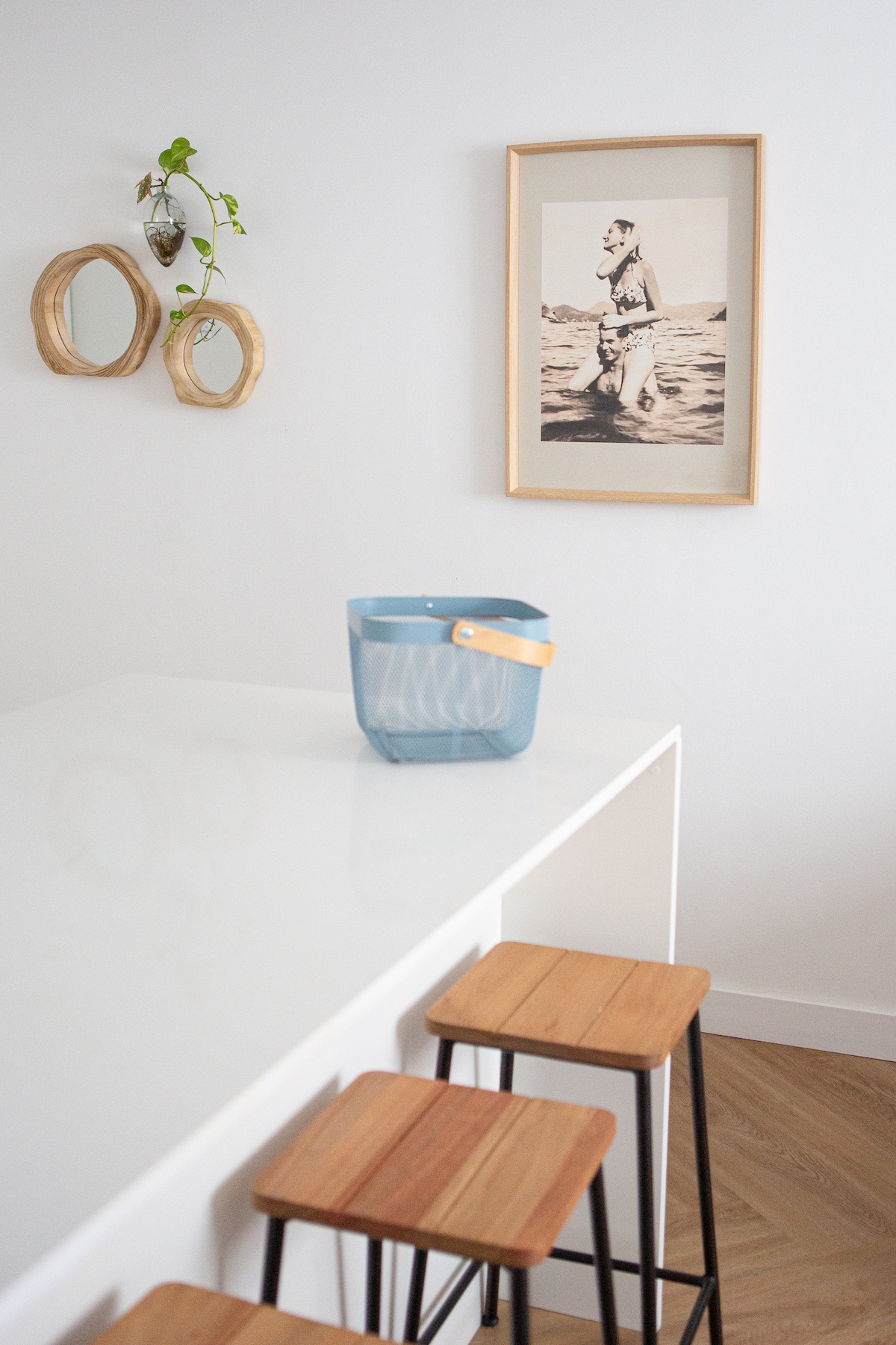 A minimalist dining area features a sleek white table topped with a blue storage basket. Three wooden stools with black legs are positioned nearby. A framed artwork and two round mirrors adorn the wall, complemented by a small plant hanging in a glass container.