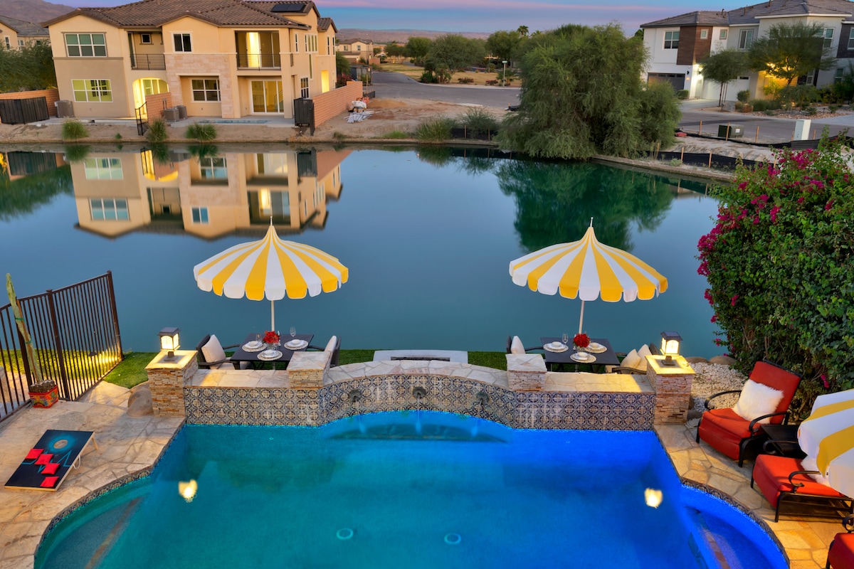 A serene outdoor area features a shimmering pool bordered by stonework. Colorful yellow and white umbrellas provide shade over lounge chairs. The calm lake reflects nearby homes and lush greenery, creating a tranquil atmosphere. Soft lighting adds to the inviting setting as dusk approaches.