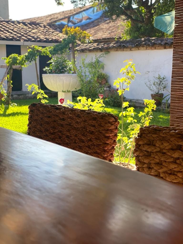 A serene view of a garden area is framed by woven chairs. Lush greenery and flowering plants enhance the natural setting, while a water fountain serves as a focal point. Sunlight filters softly through the scene, creating a tranquil and inviting atmosphere.