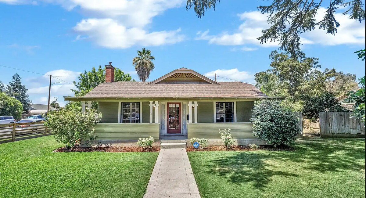 A historical single-story home is set in a green yard, with flowering shrubs and trees bordering a straight pathway leading to the entry. A covered porch is visible, featuring symmetrical windows and accents that enhance its architectural charm.