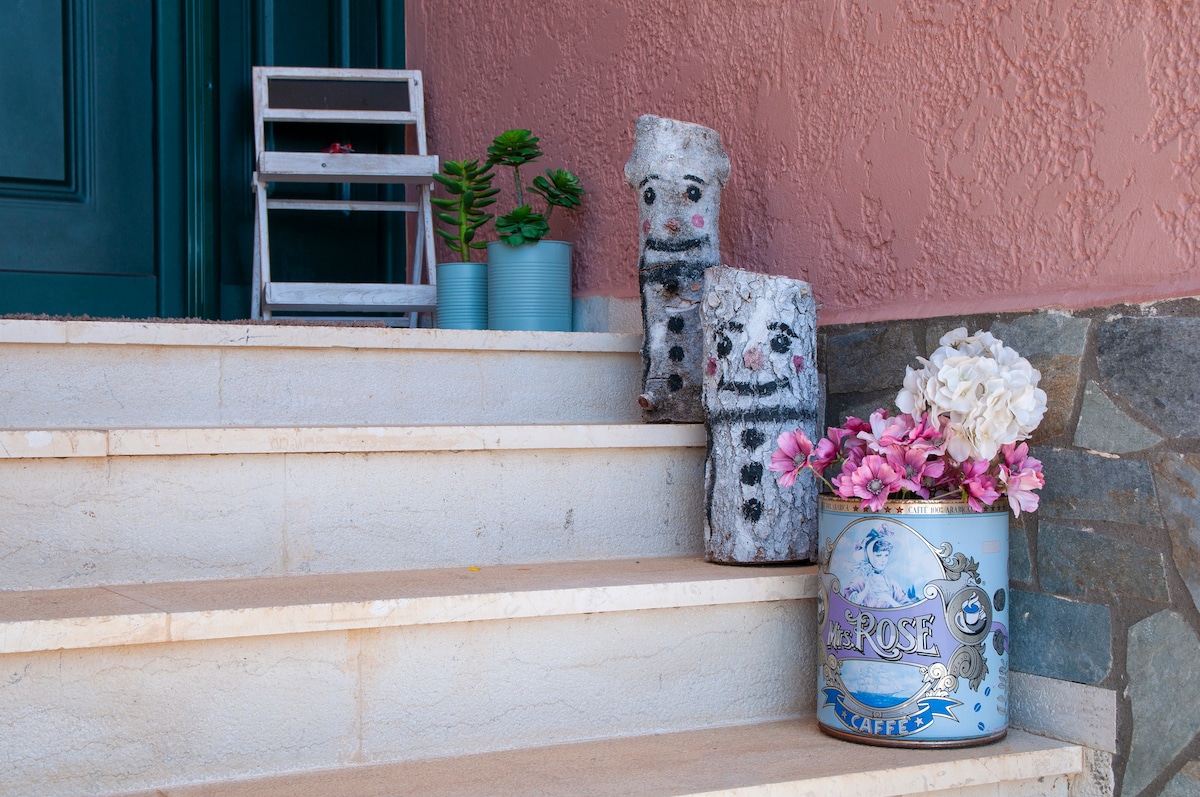 The entrance features a staircase with light-colored steps leading up to a doorway. On the steps, decorative elements include two whimsical log figures with painted faces and a blue metal container filled with pink flowers. A small potted plant adds a touch of greenery.
