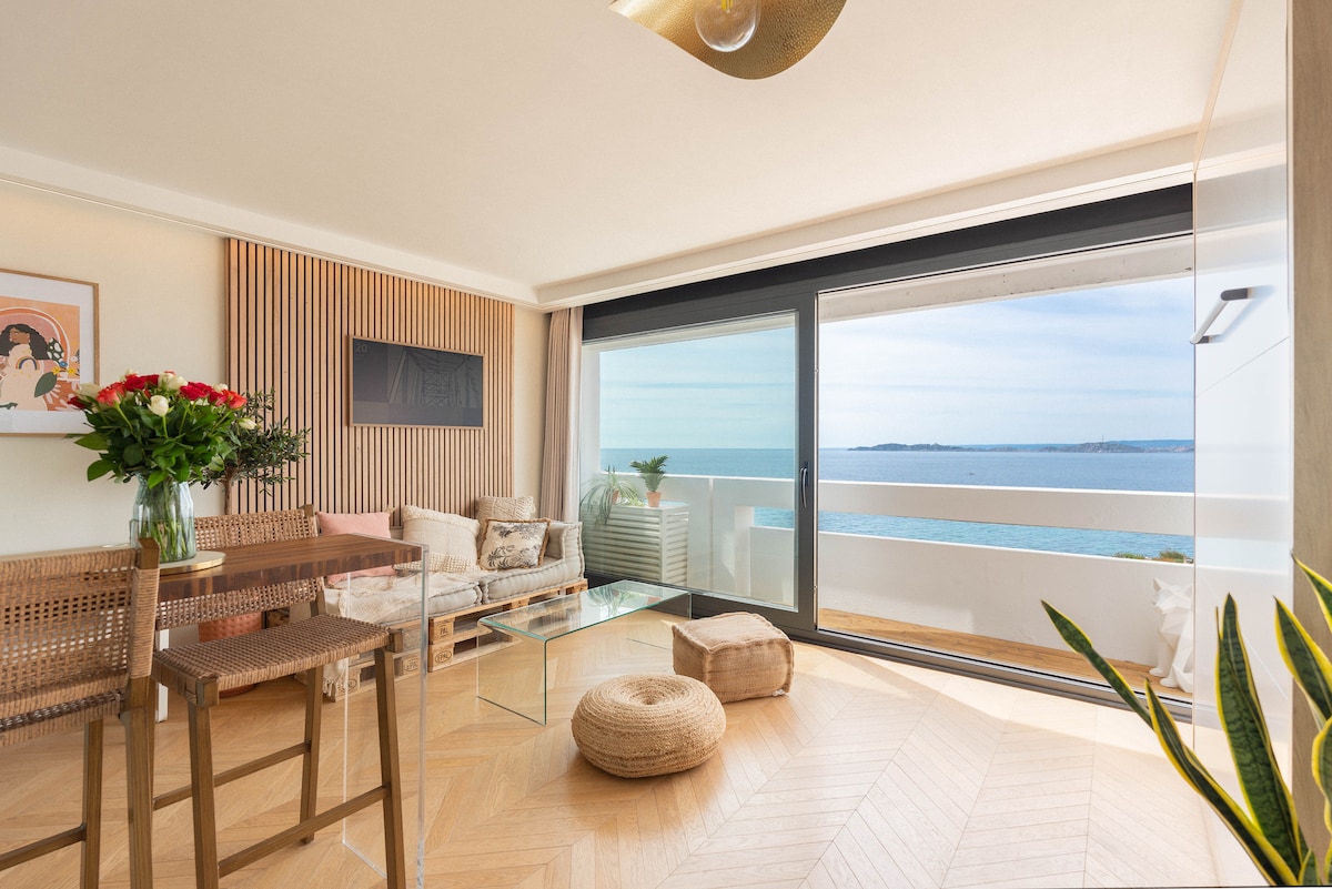 The living area features a spacious seating arrangement that incorporates natural tones. Large glass doors provide an uninterrupted view of the sea. A small dining table is positioned near a bouquet of flowers, while a pouf and decorative elements enhance the space's functionality.