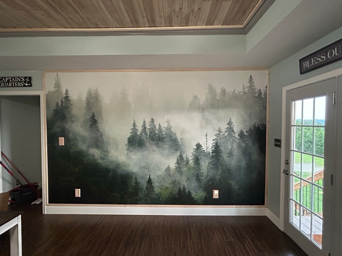 A large mural depicting a misty forest scene adorns one wall, featuring tall trees fading into a foggy background. Natural light filters through a door, adding depth to the serene atmosphere. The floor is polished and reflects the soft, wooded theme.