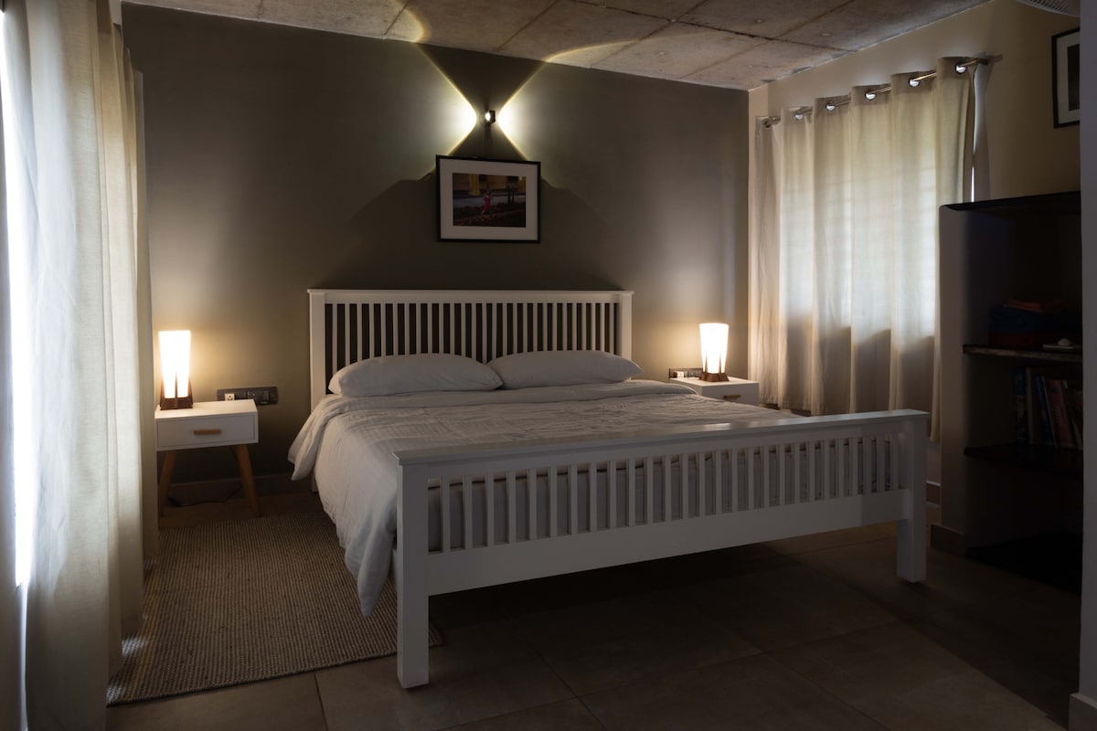 A spacious room features a large double bed with a light-colored covering, flanked by two bedside lamps illuminating the space. Soft curtains frame the windows, allowing subtle light to filter in. A shelf is visible in the corner, adding functionality to the serene atmosphere.