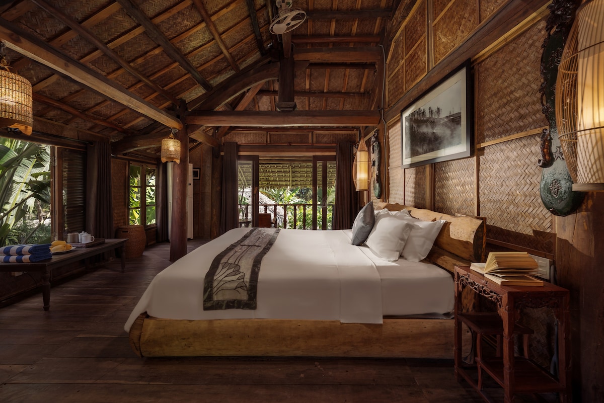 A spacious bedroom features natural wood accents and traditional woven wall decor. A large bed is positioned in the center, complemented by decorative light fixtures. Expansive windows provide views of the lush garden outside, enhancing the serene atmosphere.