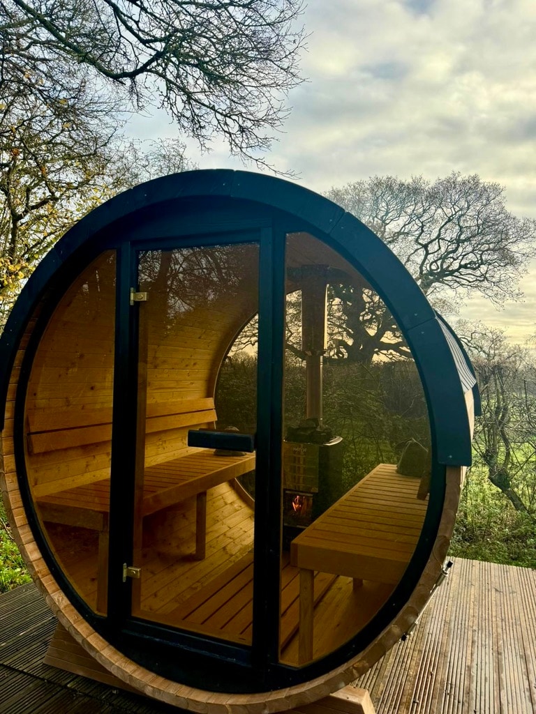 A rustic wooden sauna structure features a round design with large glass doors, inviting natural light inside. Inside, wooden benches line the interior, providing a cozy space for relaxation. The serene surroundings of trees and greenery can be seen through the glass, enhancing the tranquil atmosphere.
