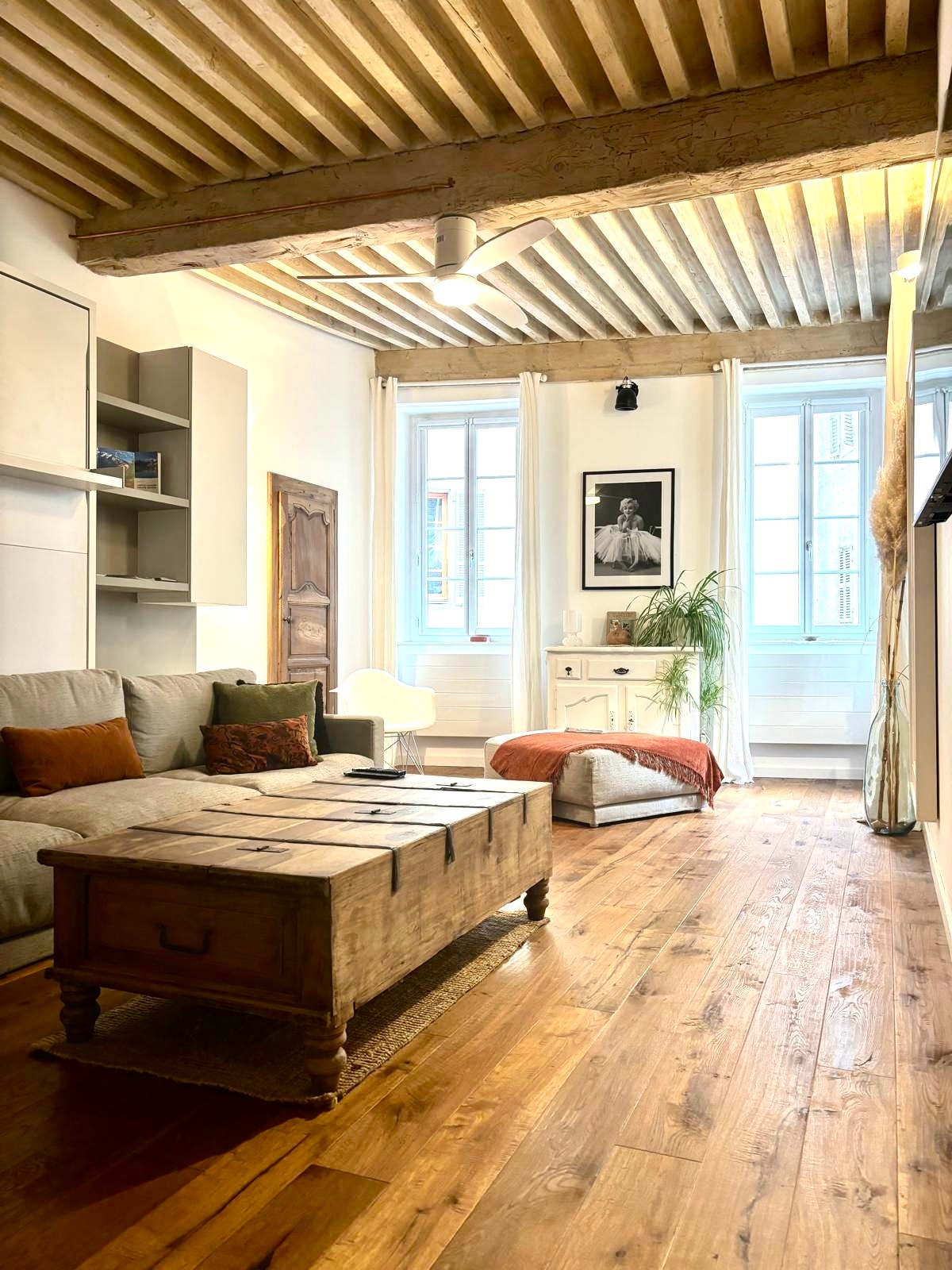 A living area is presented with wooden flooring and exposed beams overhead. A comfortable couch is paired with rustic furniture, including a coffee table and an ottoman. Natural light enters through the large windows dressed in light curtains, enhancing the space's welcoming feel.