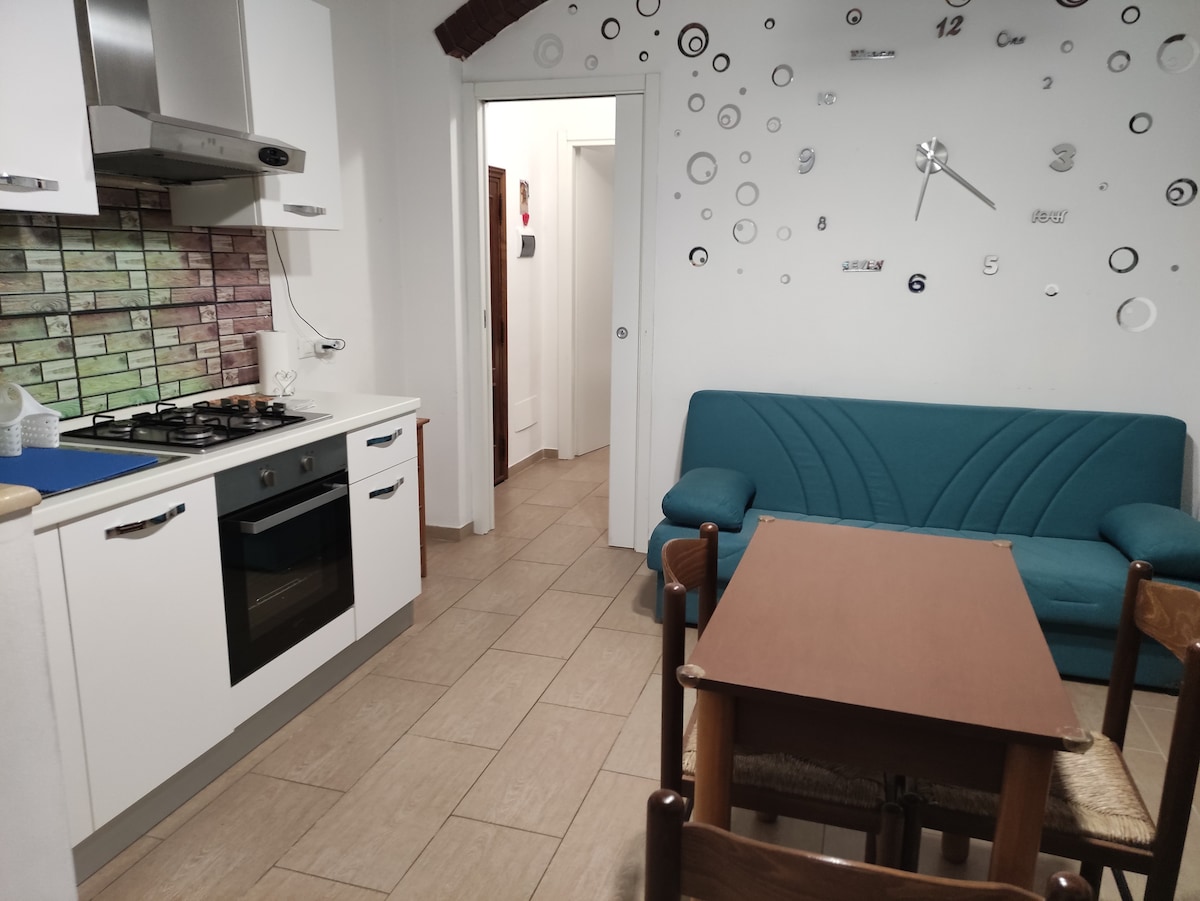 A compact kitchen area features modern appliances, including a gas stove and an oven, with a vibrant tiled backsplash. A small dining table sits adjacent to a teal sofa, which complements the light-colored walls adorned with decorative circular designs.