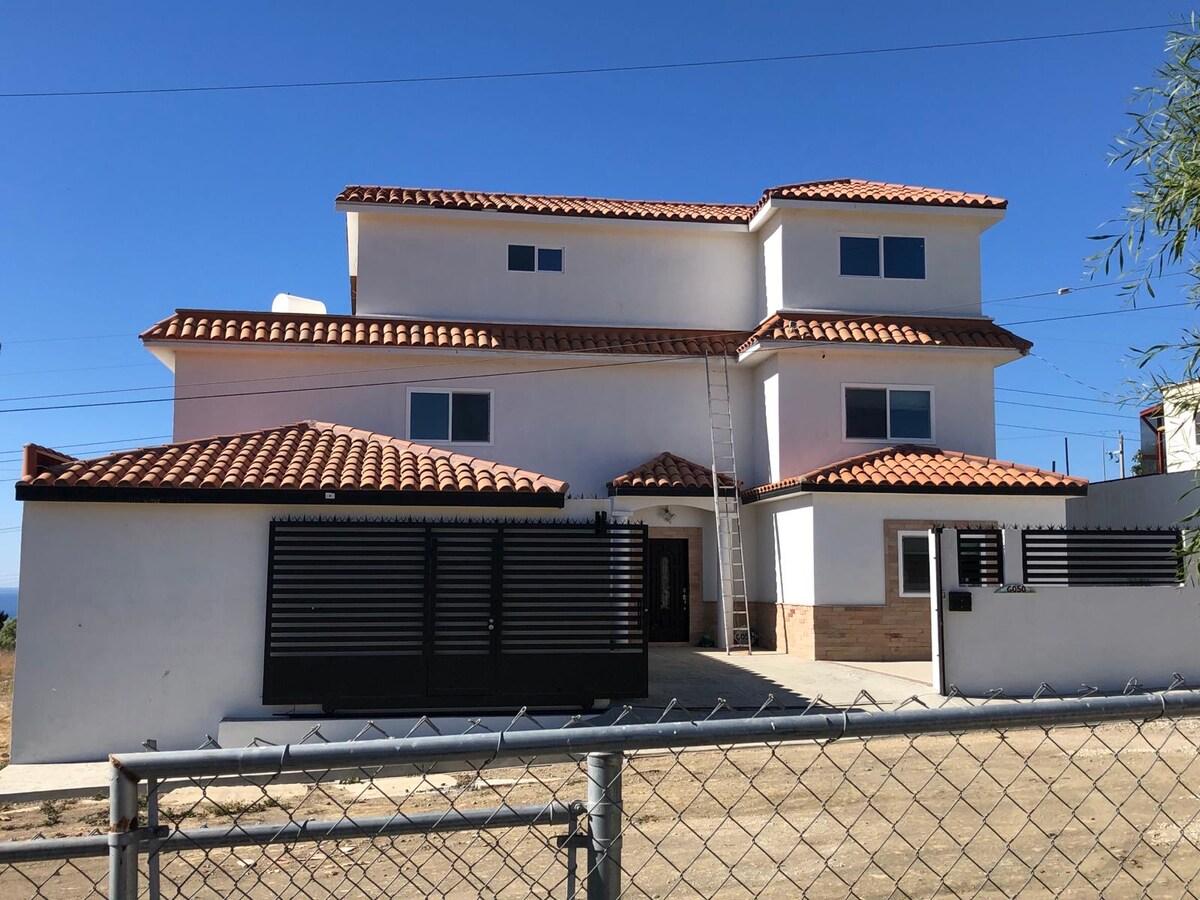 A three-story building is shown with a smooth white exterior and a terracotta roof. The facade features several large windows allowing natural light. A gated entrance in the front provides security, while a ladder leans against the side of the structure for maintenance.