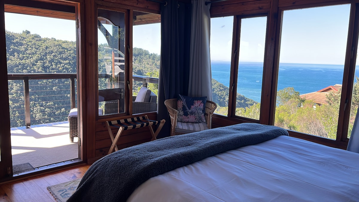 A serene bedroom is presented with large windows offering panoramic views of the ocean and surrounding greenery. A comfortable king-size bed is complemented by a cozy blanket. A seating area includes a woven chair with decorative pillows, enhancing the room's inviting nature.
